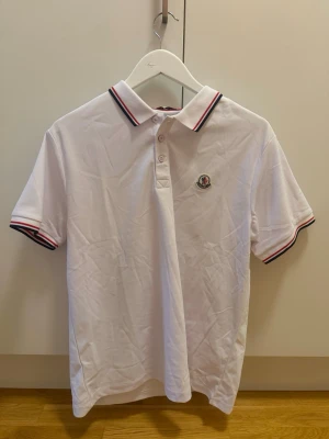 Moncler polo  - Snygg vit pikétröja med krage och knäppning framtill. Tröjan har röda och blå ränder på krage och ärmslut samt en broderad logga på bröstet. Perfekt för en stilren och sportig look.