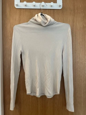 Beige ribbad polotröja från Zara - Säljer en beige ribbad polotröja från Zara. Tröjan har långa ärmar, smala ränder och en hög polokrage. Materialet är mjukt och stretchigt, perfekt för lager på lager. Enkel och stilren design som passar till det mesta.