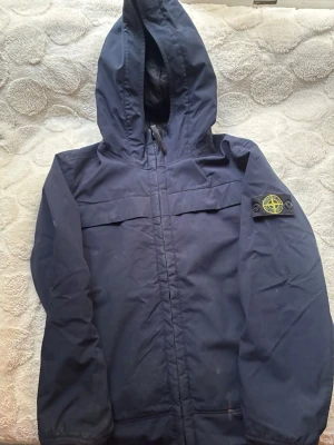 Marinblå vindjacka från Stone Island - Snygg marinblå vindjacka från Stone Island med huva och dragkedja framtill. Jackan har den klassiska logotypen på vänster ärm och är tillverkad i ett slitstarkt material. Perfekt för dig som vill ha en stilren och sportig look.