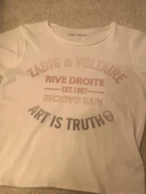 Vit Zadig & Voltaire t-shirt - Vit t-shirt från Zadig & Voltaire med tryck i rött och svart framtill. Texten 'RIVE DROITE', 'ART IS TRUTH' och en liten döskalle ger en cool vibe. Klassisk rund hals och korta ärmar. Perfekt till jeans eller shorts.