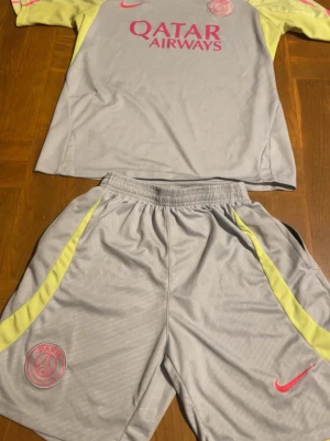 PSG grått matchställ med gula detaljer - Säljer ett PSG matchställ från Nike i ljusgrått med gula detaljer på ärmar och shorts. Tröjan har rosa PSG-logga och Qatar Airways-tryck framtill, samt rosa Nike-logga. Shortsen har elastisk midja och PSG-märke. Perfekt för fotbollsträning eller match. Passar Både M och L