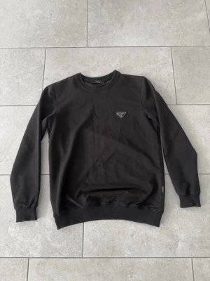 Svart sweatshirt från Prada - Stilren svart sweatshirt från Prada med rund halsringning och ribbade muddar. Tröjan har en ikonisk trekantig Prada-logga på bröstet och är tillverkad i mjukt bomullsmaterial. Perfekt för en clean och trendig look.