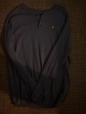 Blå långärmad lyle&scott - Mysig blå långärmad tröja med rund halsringning och knappdetalj framtill. Tröjan har en gul broderad logga på bröstet och är tillverkad i ett mjukt material som passar perfekt för kyligare dagar. Vintage stil