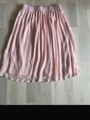 Ljusrosa plisserad kjol från Lindex - Söt ljusrosa plisserad kjol från Lindex med bred resår i midjan. Kjolen har ett luftigt och flowy tyg som ger en fin siluett. Perfekt för dig som gillar en feminin och enkel stil.