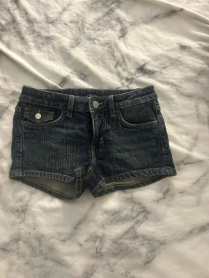 Mörkblå denimshorts med fickor - Snygga mörkblå denimshorts med klassisk femficksdesign och silverfärgade knappar. Shortsen har bälteshällor och korta ben, perfekta för varma dagar. Passar dig som gillar en avslappnad och trendig stil.