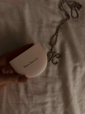 Rare Beauty kompakt powder - Snygg kompakt foundation från Rare Beauty i en ljusbeige nyans. Kommer i ett stilrent, halvmåneformat etui i ljusrosa plast med spegel och separat fack för svamp. Perfekt att ha med i väskan för touch-ups under dagen.