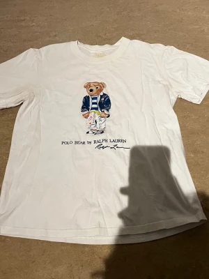 Vit Polo Bear t-shirt från Ralph Lauren - Vit t-shirt med Polo Bear-tryck från Ralph Lauren. Klassisk passform med korta ärmar och rund halsringning. Trycket visar en björn i sportig outfit och texten 'Polo Bear by Ralph Lauren' under. Perfekt för en avslappnad och trendig look.