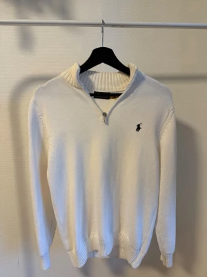 Vit stickad tröja från Ralph Lauren - Vit stickad tröja från Ralph Lauren med halv dragkedja och broderad logga på bröstet. Tröjan har ribbstickad krage, ärmslut och nederkant. Perfekt för en stilren och klassisk look.