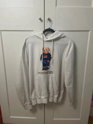 Polo Ralph Lauren Bear Hoodie - Säljer en vit RL hoodie med huva och dragsnören. På framsidan finns ett stort tryck av en björn i jeans och blå tröja. Hoodien har magficka och ribbade muddar vid ärmar och nederkant. Perfekt för en chill och avslappnad stil.