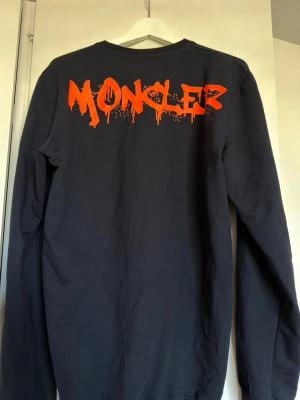 Svart långärmad Moncler t-shirt - Svart långärmad t-shirt från Moncler med stora orange tryck på ryggen och logopatch på bröstet och ärmen. T-shirten har en avslappnad passform och är tillverkad i mjuk bomull. Perfekt för dig som gillar streetwear och vill sticka ut.