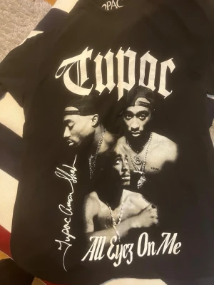 Svart Tupac t-shirt med tryck - Säljer en svart t-shirt med stort Tupac-tryck på framsidan och texten 'All Eyez On Me'. T-shirten har korta ärmar och är gjord i mjuk bomull. Perfekt för dig som gillar hiphop och streetwear.