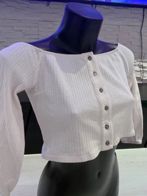 Vit croppad blus med knappar - Trendig vit croppad blus med långa ärmar och snygga knappar framtill. Blusen är ribbad och har en offshoulder-design som ger en modern och avslappnad look. Perfekt att matcha med höga jeans eller kjol.