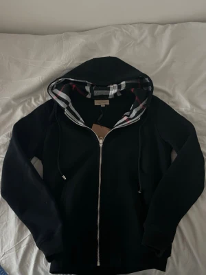 Svart hoodie med dragkedja och huva - Säljer en svart hoodie med dragkedja och huva. Insidan av huvan har ett klassiskt rutigt mönster i vitt, svart och rött. Hoodien har dragsko i huvan och ribbade muddar vid ärmar och nederkant. Perfekt för en chill och stilren look.
