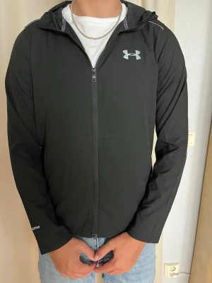 Svart vindjacka från Under Armour - Storlek M, passar verkligen S! Snygg svart vindjacka från Under Armour med huva och dragkedja framtill. Jackan har logga på bröstet och stor text på ryggen. Tillverkad i lätt och tunt material, perfekt för blåsiga dagar. Ventilationspanel på ryggen för extra komfort.