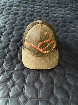 Gucci keps med orm och GG-mönster - Snygg keps från Gucci med klassiskt GG-mönster i brunt och svart mesh på sidorna. Framtill pryds kepsen av en röd och vit orm som ger en unik och trendig look. Perfekt accessoar för dig som vill sticka ut och addera lite lyx till outfiten.