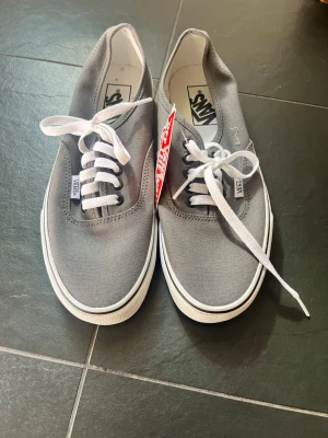 Gråa Vans sneakers med vita detaljer - Snygga gråa Vans sneakers i klassisk låg modell med vita snören och vit sula. Skorna har canvasmaterial och Vans-logga på sidan samt innersulan. Perfekta för en clean och stilren look.