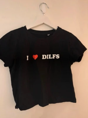 Svart t-shirt med text I ❤️ DILFS - Svart t-shirt i bomull med vit text och ett rött hjärta på bröstet där det står 'I ❤️ DILFS'. Klassisk rund halsringning och korta ärmar. Perfekt för dig som vill ha en statement-tee med humor.