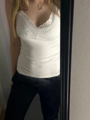 Vit ribbad linnetopp med spets - Säljer en vit ribbad linnetopp med v-ringning och fina spetsdetaljer vid halsen. Toppen är ärmlös och har en slim passform som sitter snyggt på kroppen. Perfekt att styla med jeans eller kjol för en trendig look.