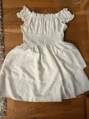 Vit offshoulder kortklänning - Söt vit kortklänning med offshoulder och volangdetaljer. Klänningen har smock i midjan för en bekväm passform och ett diskret prickigt mönster i tyget. Perfekt för varma dagar och somriga tillfällen.