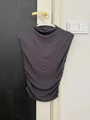 Svart ärmlös polotopp - Säljer en svart ärmlös polotopp med hög hals och lätt rynkade sidor. Toppen har en clean och stilren look som passar perfekt till jeans eller kjol. Materialet känns mjukt och stretchigt, troligen bomull eller polyester.