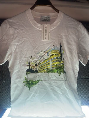 Vit Casablanca t-shirt med tryck - Säljer en vit t-shirt från Casablanca med ett stort färgglatt tryck framtill som föreställer en byggnad och palmer. T-shirten har rund hals och korta ärmar. Perfekt för dig som gillar snygga statement-plagg.