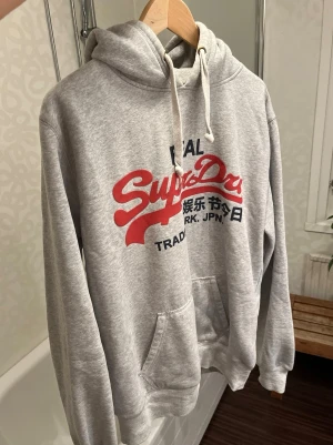Grå Superdry hoodie med tryck - Säljer en grå hoodie från Superdry i mycket fint skick. Jag är 189cm. Kontakta vid frågor!