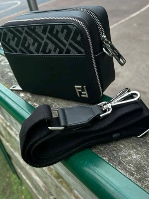 FF Väska - Snygg svart axelväska med silverfärgade detaljer och dubbla dragkedjor. Väskan har ett tydligt FF-mönster i grått och svart på framsidan och baksidan samt en bred, justerbar axelrem. Tillverkad i en mix av läder och textilmaterial.