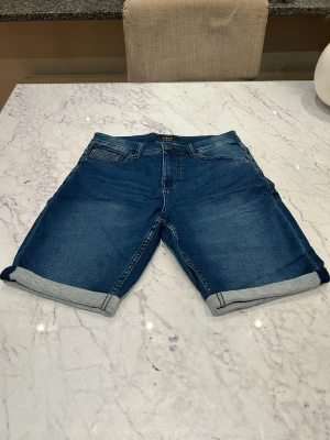 Blå jeansshorts med uppvik - Snygga blå jeansshorts med klassisk femficksdesign och uppvikta benslut. Shortsen har normal passform och stängs med dragkedja och knapp. Perfekta för varma dagar och enkla att matcha med olika stilar.