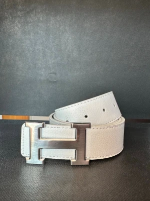 Vitt skärp med Silver H-spänne - Snyggt vitt skärp i skinn med en stor, silverfärgad H-formad metallspänne. Bältet har en stilren och modern look med synliga sömmar och är vändbart med svart insida. Perfekt för att lyfta vilken outfit som helst.