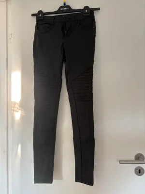 Svarta bikerjeans med ribbade detaljer - Snygga svarta bikerjeans med tight passform och ribbade detaljer över knäna. Jeansen har klassisk femficksdesign och dragkedja med knapp. Perfekta för dig som gillar en edgy och modern stil. Storlek M bra skick!