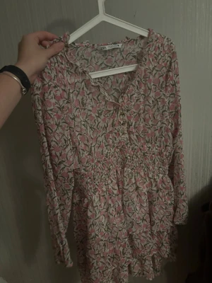 Blommig kortklänning med knappar - Söt kortklänning med rosa och grönt blommönster på vit botten. Klänningen har långa ärmar, smock i midjan och dekorativa knappar framtill. Tillverkad i ett lätt och luftigt tyg, perfekt för varmare dagar. Liten i storleken, passar en S/M