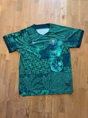 Nike Nigeria 2022/23 Stadium Home Jersey – Storlek L - 100 % autentisk✅. Tröjan är helt ny och aldrig använd, i nyskick utan några defekter, fläckar, repor eller andra skador. Tillverkad med Nikes Dri-FIT-teknologi som ger hög komfort och god andningsförmåga – perfekt för både match, träning, vardagsbruk eller som ett samlarobjekt.  📦 Snabb och säker leverans. • Märke: Nike • Lag: Nigeria • Modell: Stadium Home Jersey 2022/23 • Storlek: L • Färg: Grön/Vit • Skick: Helt ny – aldrig använd