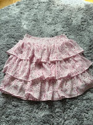 Blommig volangkjol från FB Sister - Supersöt kort kjol från FB Sister med volanger och blommigt mönster i rosa och vitt. Kjolen har resår i midjan och flera lager volanger som ger en fluffig och lekfull look. Perfekt för dig som gillar romantisk och trendig stil.