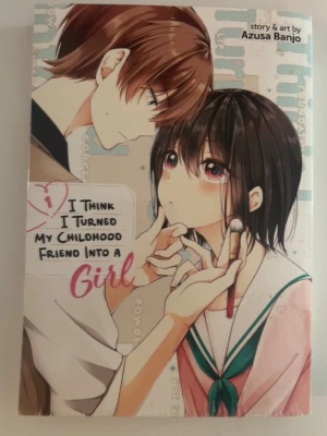 I Think I Turned My Childhood Friend - Manga med färgglad omslagsbild som visar två tonåringar, en med kort brunt hår och en med svart hår, där den ena applicerar smink på den andra. Serien är skapad av Azusa Banjo och handlar om vänskap, förvandling och romantik med humoristisk twist.