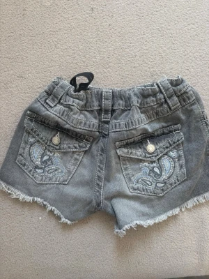 Grå jeansshorts från Gina Tricot - Säljer ett par grå jeansshorts från Gina Tricot med fransig kant och snygga broderade detaljer på bakfickorna. Shortsen har normal passform och elastisk midja för extra komfort. Perfekta för varma dagar och enkel att matcha med olika toppar.