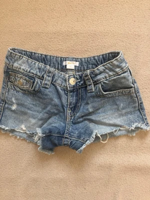 Blå jeansshorts från Gina Tricot - Säljer ett par blå jeansshorts från Gina Tricot med råa kanter och slitna detaljer. Klassisk femficksmodell med knapp och dragkedja framtill. Perfekta för varma dagar och en avslappnad stil.