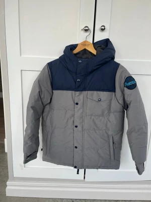 Grå och marinblå vinterjacka  - Vinter jacka från Burton i grått med marinblått axelparti och huva. Jackan har en bröstficka med knapp, två sidofickor och en stor Burton-logga på ärmen. Tjock vaddering och sportig look, perfekt för vintern.