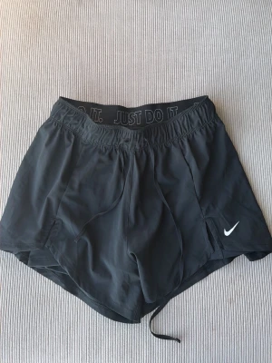 Svarta löparshorts från Nike - Snygga svarta löparshorts från Nike med elastisk midja och snörning. Diskret Nike-logga på benet och texten 'JUST DO IT.' på insidan av linningen. Tillverkade i lätt och snabbtorkande material, perfekta för träning och löpning.