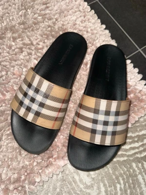 Burberry rutiga slides sandaler - Säljer ett par klassiska slides från Burberry med det ikoniska beige, svarta, vita och röda rutmönstret. Sandalerna har svart sula i gummi och bred rem i textil med Burberrys signaturmönster. Perfekta för en trendig och avslappnad stil.