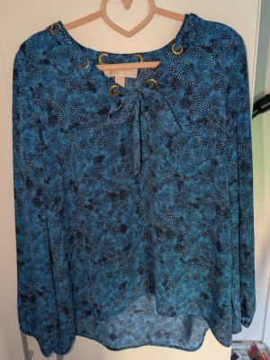 Blå mönstrad blus med knytning - Blå blus med abstrakt prickigt mönster i svart och vitt, närmare bild på mönstret finns på bild 3.. Blusen har långärmad design, rund halsringning med dekorativa öljetter och knytband framtill. Tillverkad i ett lätt och luftigt material som ger en avslappnad look.