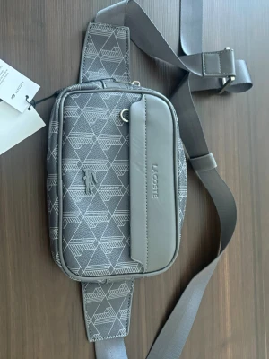 Grå magväska från Lacoste - Snygg grå magväska från Lacoste med geometriskt mönster och justerbart band. Väskan har två fack med dragkedja och en präglad Lacoste-logga på framsidan. Tillverkad i slitstarkt syntetmaterial och passar perfekt för att bära det viktigaste.