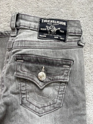 Grå True Religion jeans - Säljer ett par grå jeans från True Religion med ikoniska ficklock och kontrastsömmar. Jeansen är i ett jätte bra skick. Inga hål eller något annat som har förstört dom! Dom är använda i ca ett halvår. Dom har tyvärr blivit korta nu! Pris kan diskuteras💕💐