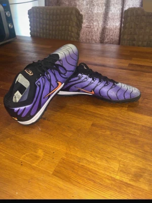 Lila och svarta Nike fotbollsskor - Säljer ett par snygga fotbollsskor från Nike i lila och svart med orange swoosh. Skorna har en låg profil, snörning och mönstrad sula för bra grepp på konstgräs. Materialet är syntet med meshdetaljer för bra ventilation. Perfekta för dig som vill sticka ut på planen!