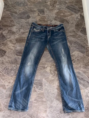 Blå bootcut jeans med slitningar - Säljer ett par blå secondhand bootcut jeans med snygga slitningar och kontrastsömmar. Jeansen har broderade bakfickor och en avslappnad passform. Perfekta för en casual look och passar till det mesta. Materialet är klassisk denim. Jeansen är från rock revival. Passar en som har storlek 38 i damstorlek.