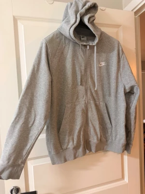 Grå zip hoodie från Nike - Snygg grå zip hoodie från Nike med vit logga på bröstet. Hoodien har dragkedja, två stora fickor framtill och justerbar huva med snören. Tillverkad i mjukt sweatshirtmaterial som är perfekt för chill eller träning.