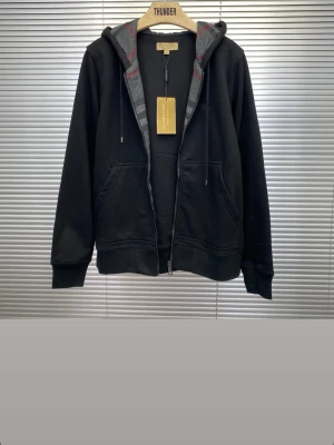Burberry kofta  - Svart burberry hoodie med dragkedja och huva, fodrad med grått och rött rutmönster i luvan. Klassisk modell med fickor framtill och justerbara snören i luvan. Perfekt för en avslappnad och stilren look. Nytt skick!