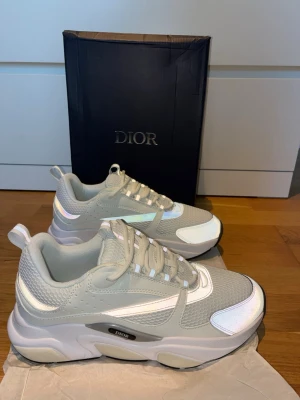 D i o r B22 sneakers  - Snygga chunky sneakers från D i o r i ljusgrått med vita och reflekterande detaljer. Ovandelen är i mesh och läder, med tjock sula och logga på sidan. Perfekta för dig som gillar en modern och sportig look.