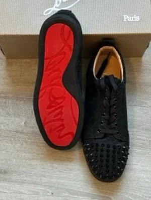 Christian Louboutin  - Snygga svarta sneakers från Christian Louboutin med ikonisk röd sula och svarta nitar på tån. Skorna har en låg profil, svarta skosnören och är tillverkade i mocka och läder. Perfekta för dig som vill sticka ut med en edgy och exklusiv stil.