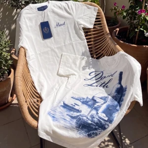 Vit t-shirt med blått tryck - Säljer en vit t-shirt med blått tryck framtill och stort motiv på ryggen. Framsidan har en liten text och baksidan visar ett konstnärligt motiv med natur och vinflaska. Klassisk passform och rund hals. Perfekt för dig som gillar stilrena men unika plagg.
