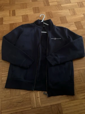 Marinblå ziptröja från Sail Racing - Snygg marinblå ziptröja från Sail Racing med hög krage och broderad logga på bröstet. Tröjan har långa ärmar, muddar vid ärmslut och nederkant samt mjukt fleecefoder på insidan. Perfekt för lager-på-lager och chill dagar.